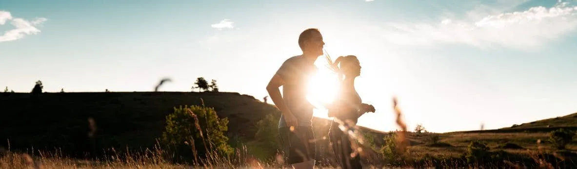 A silhouette of two people running in nature with the sun shining between them.