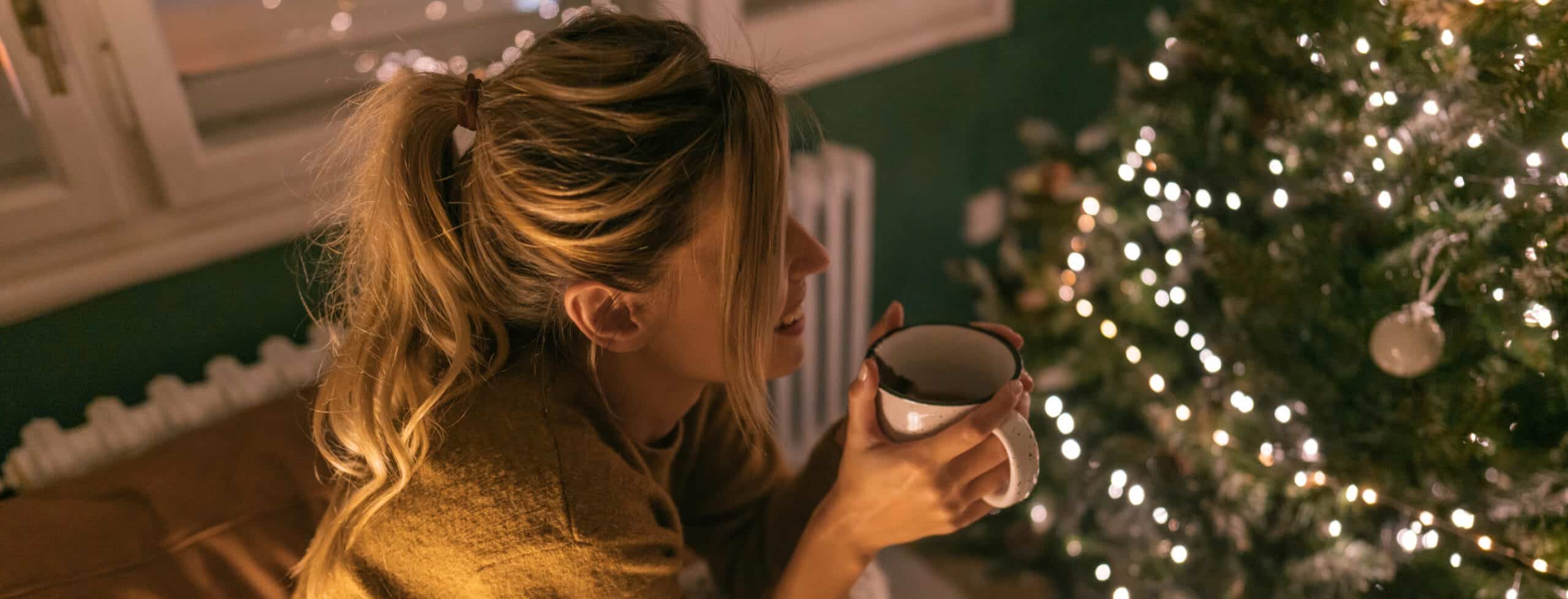 A woman drinking a cup of coffee with holiday lights and decorations behind her.