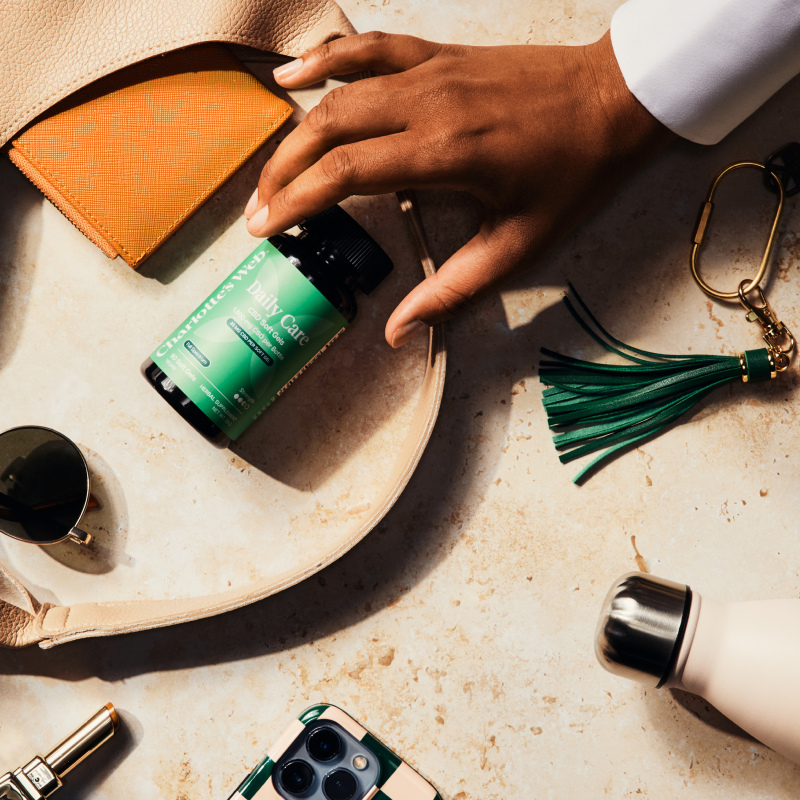 Hand holding a green bottle with a beige bag and accessories on a textured surface