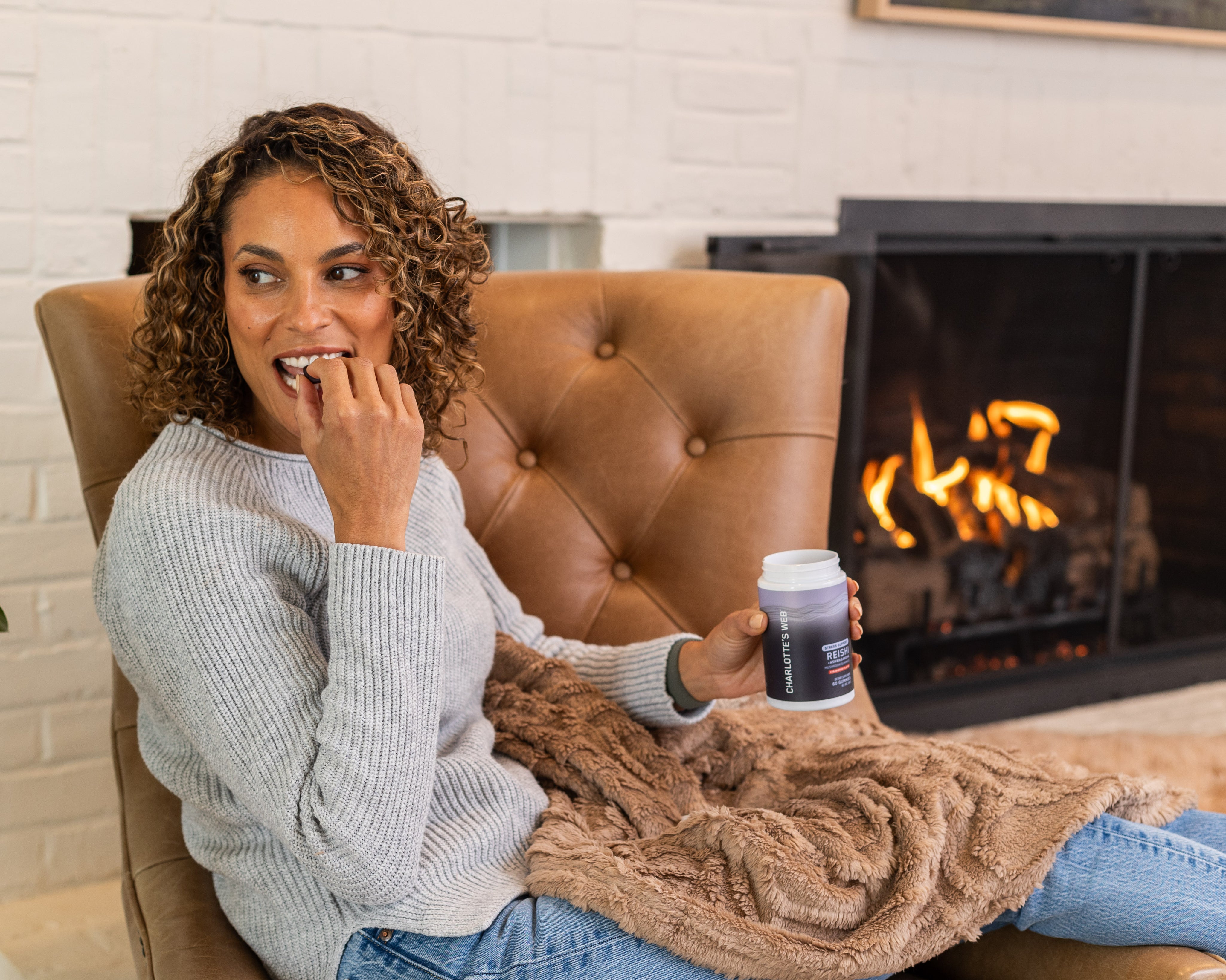 Smiling woman relaxing in a cozy chair by the fireplace, enjoying Charlotte's Web™ Stress Support gummies.