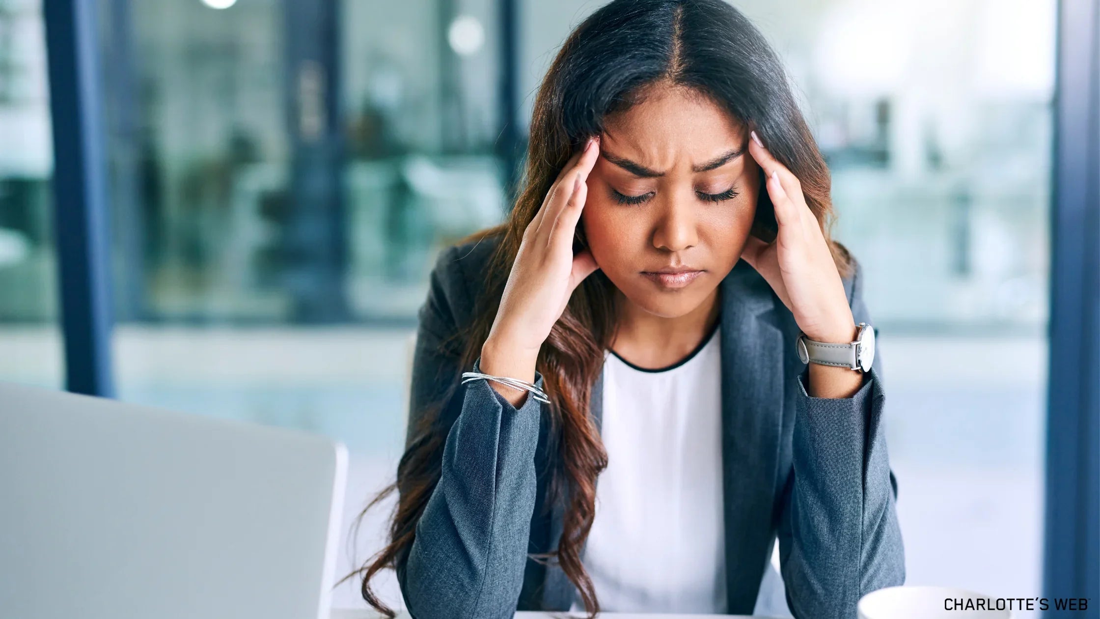 A woman in a business suit sitting at a desk, holding her head with both hands in a gesture of stress or headache. She appears overwhelmed, highlighting the need for stress management techniques.