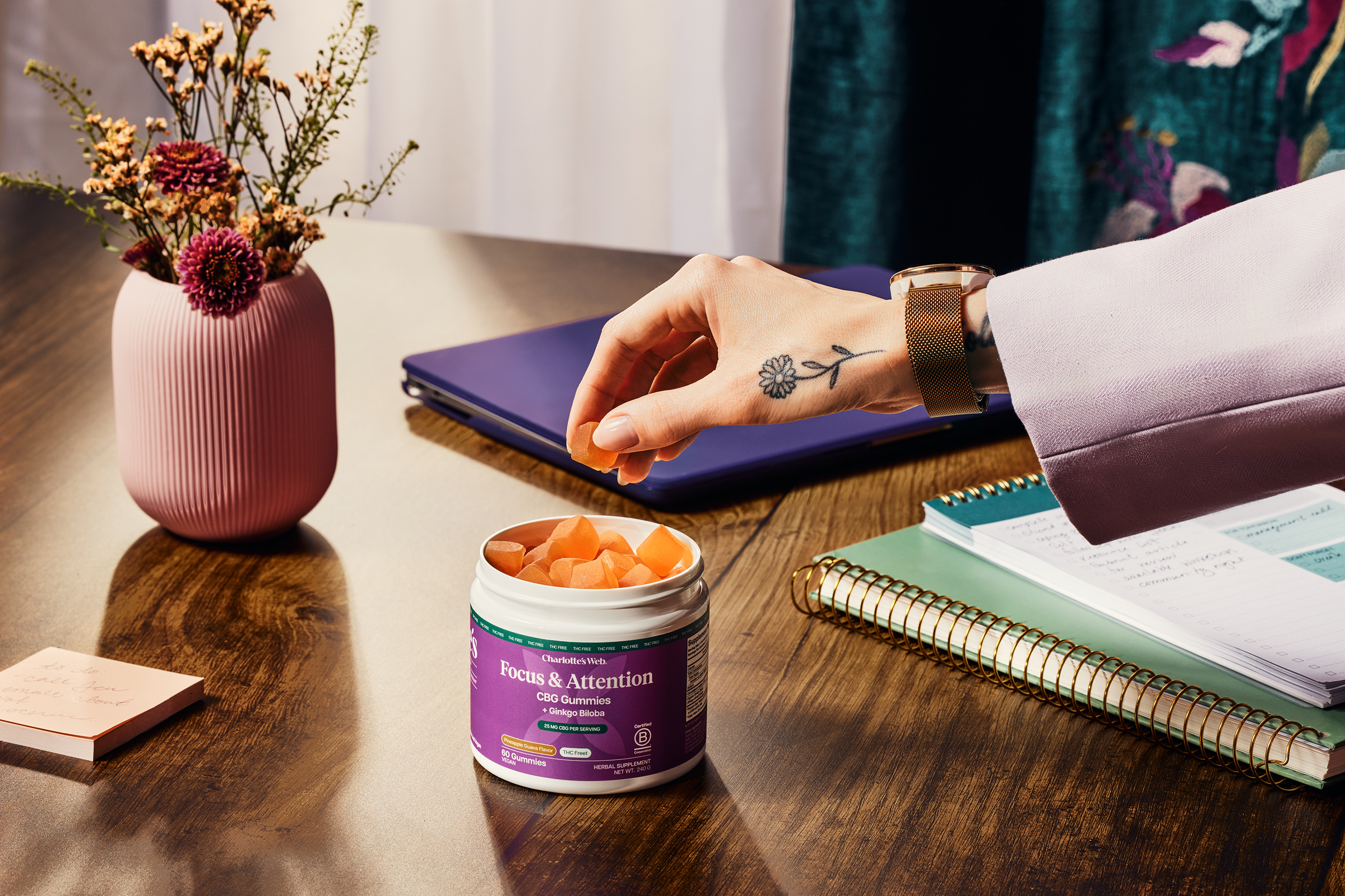 Person reaching for a Focus & Attention gummies container on a desk with a notebook and flowers in the background