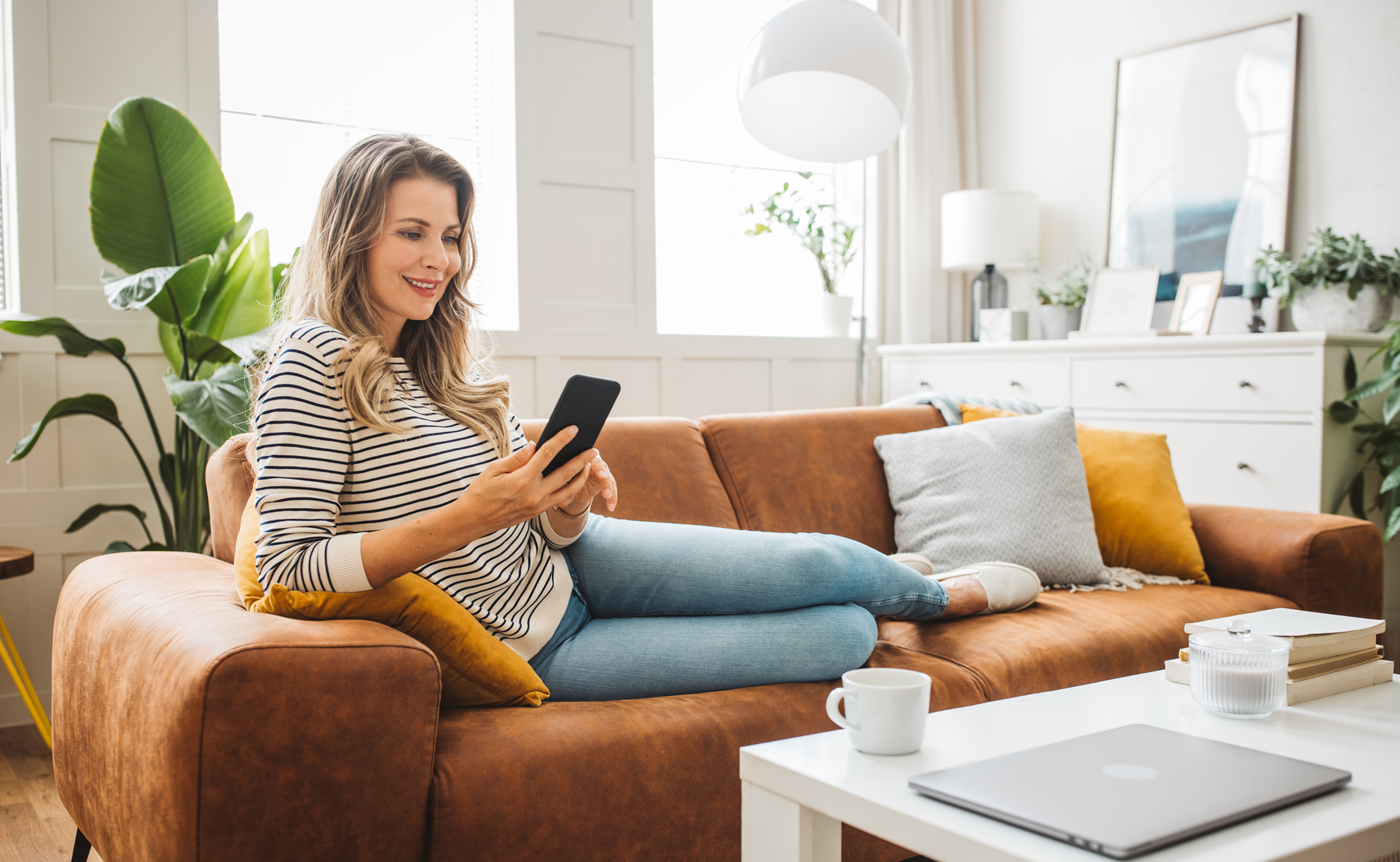 Woman sitting on a brown couch using a smartphone in a bright living room.