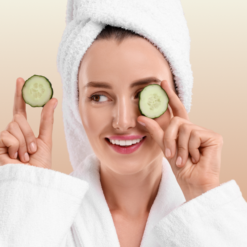 Woman in a white robe with cucumbers on her eyes against a beige background