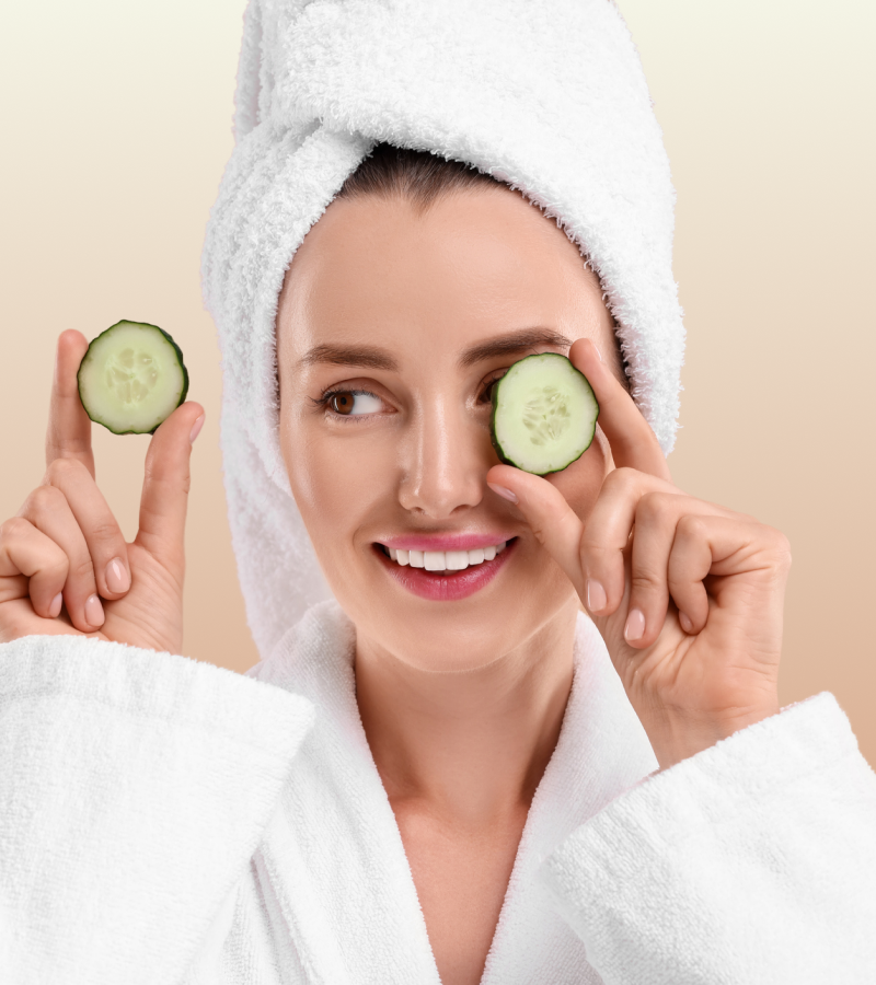 Woman in a white robe with cucumbers on her eyes against a beige background