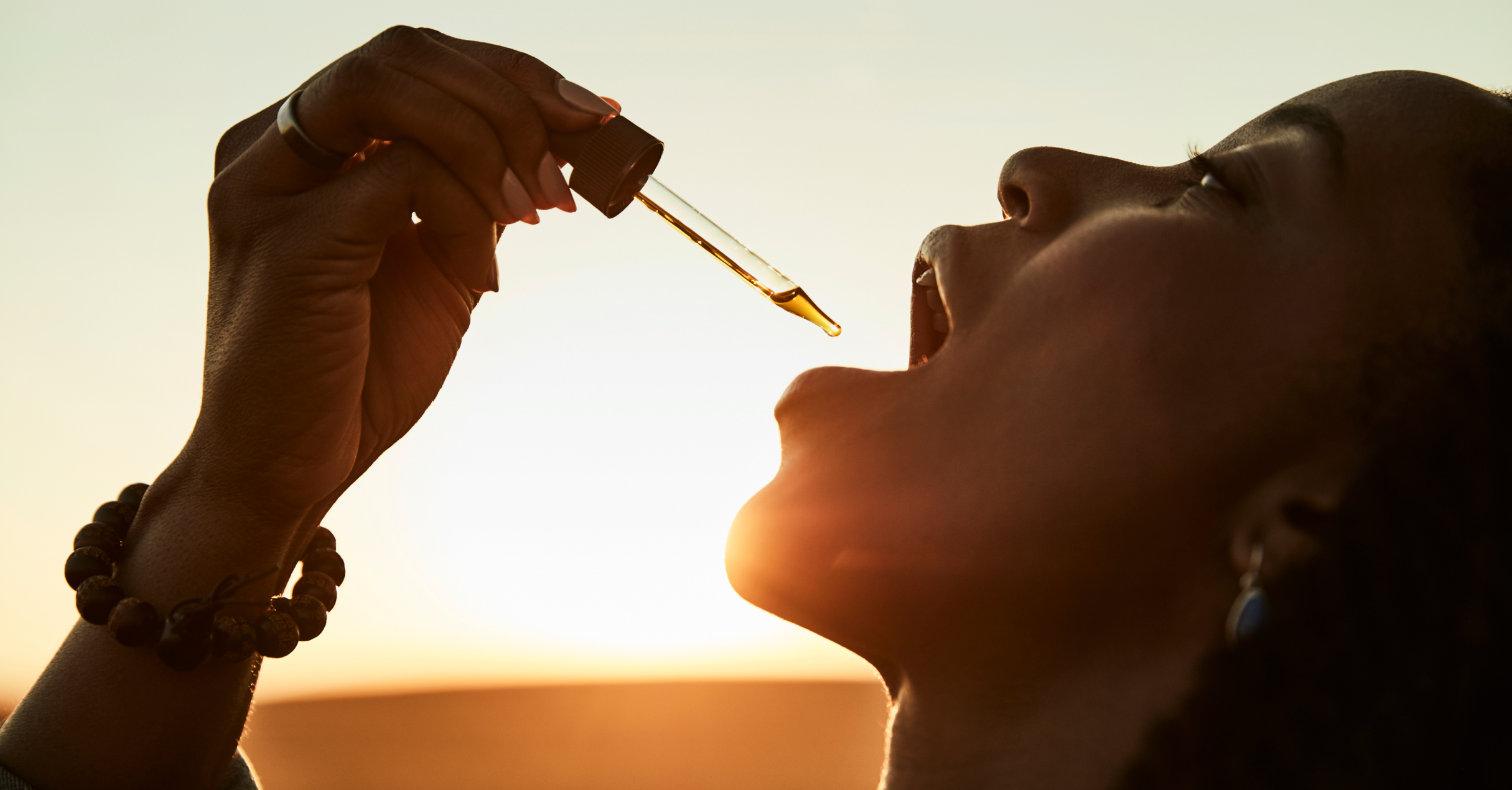 Person applying a dropper of liquid in their mouth against a sunset background