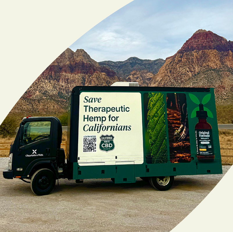 Charlotte's Web delivery truck parked outdoors beside a desert road with red rock mountains in the background. The side of the truck displays a large white panel with the text 'Save Therapeutic Hemp for Californians' and a QR code.