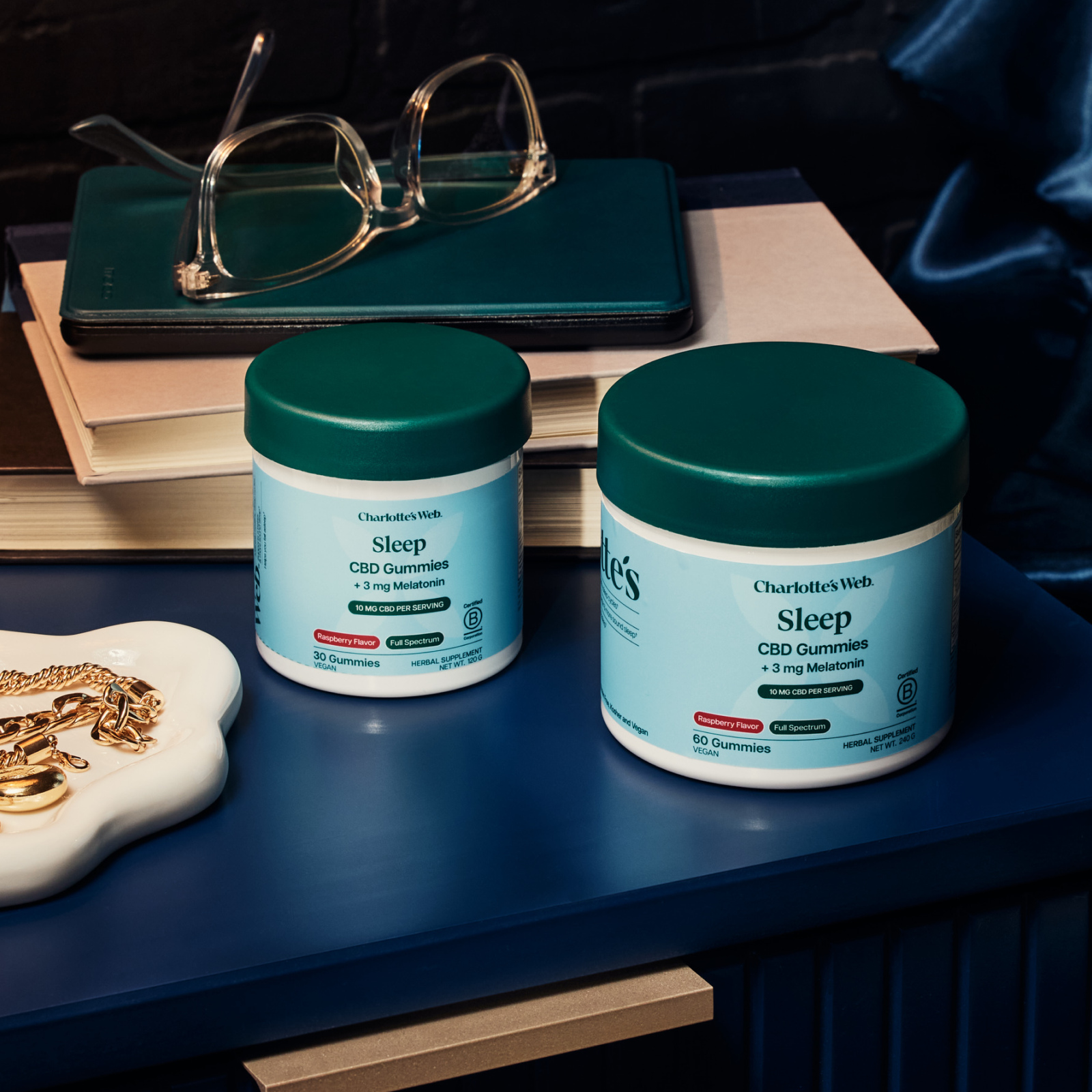 Two jars of Charlotte's Web Sleep CBD Gummies on a nightstand with books and glasses.