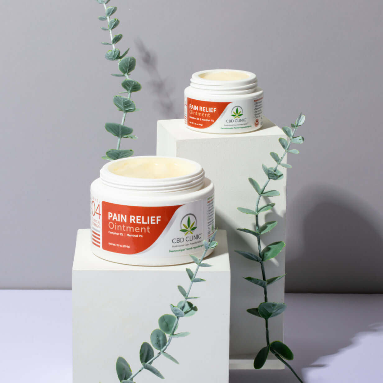 Two jars of CBD Clinic Pain Relief Ointment on white stands with green leaves on a gray background